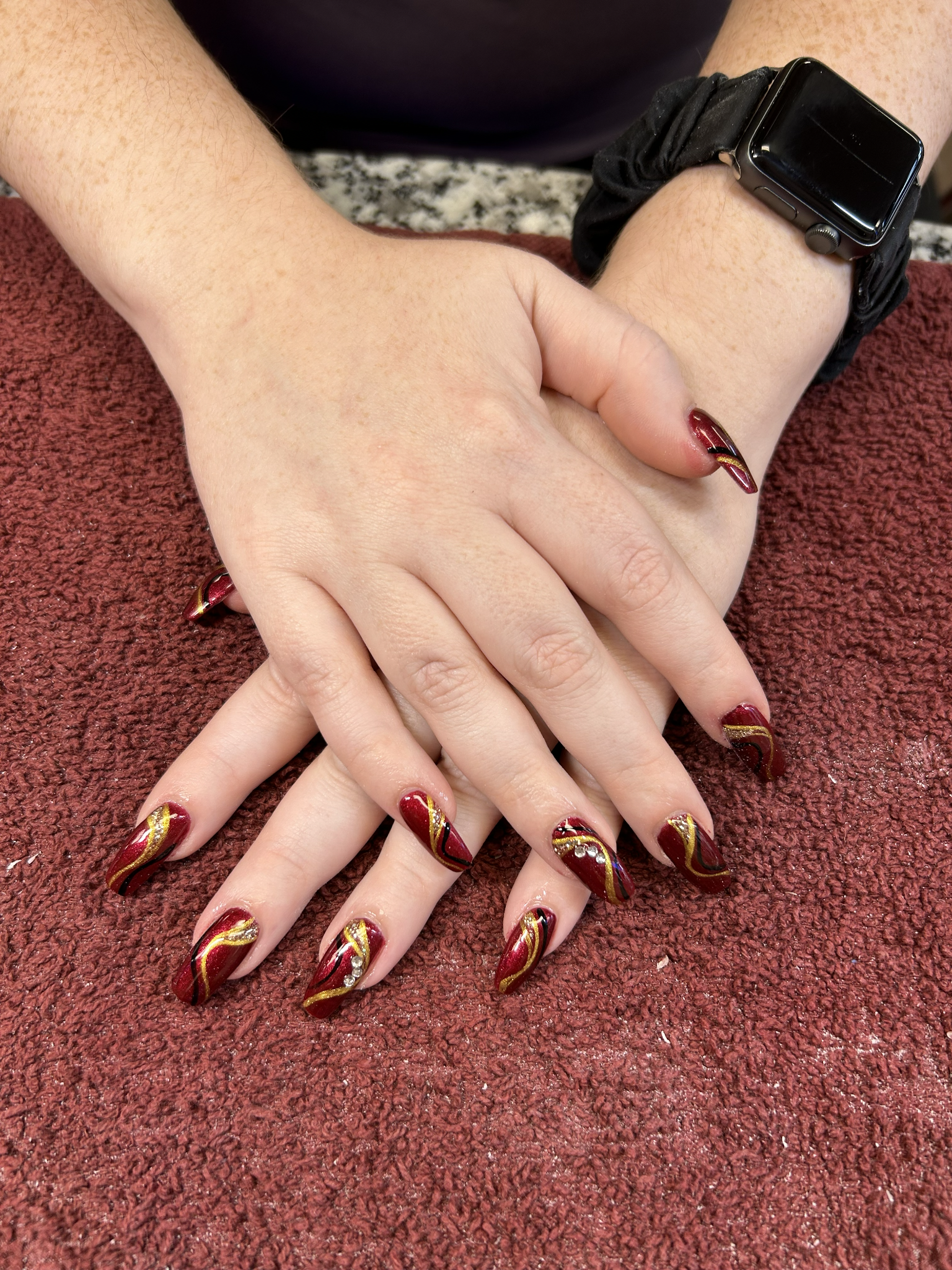 Burgundy glitter nail design with flowing gold accents and rhinestone details by Velour Nails Studio